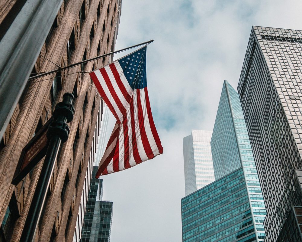 A low angle shot of the USA or United States of America flag on a flagpole near skyscrapers under a cloudy sky